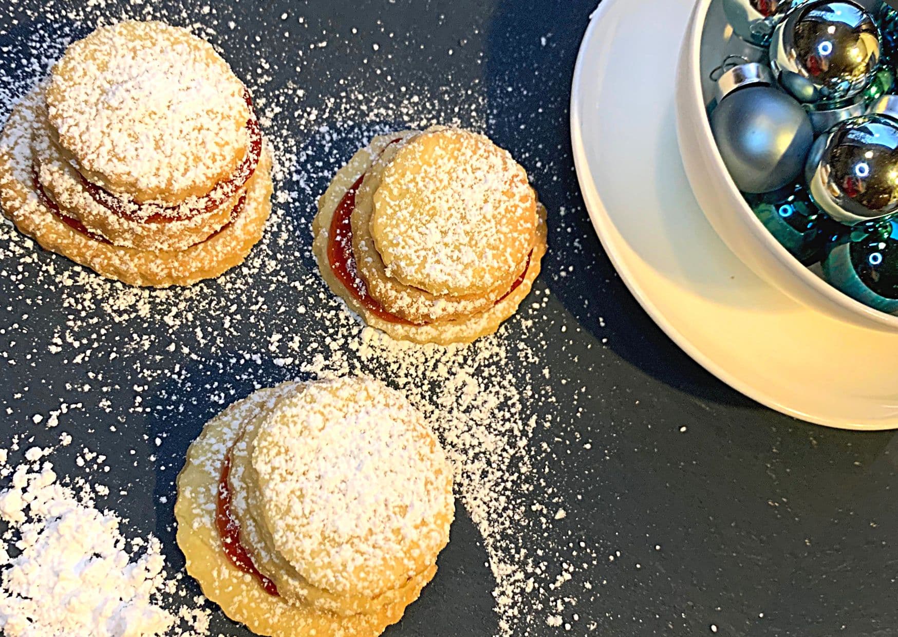 A trio of German Christmas terraces, my favourite German Christmas cookies, dusted with powdered sugar, on a blue background with a white bowl of blue Christmas baubels.