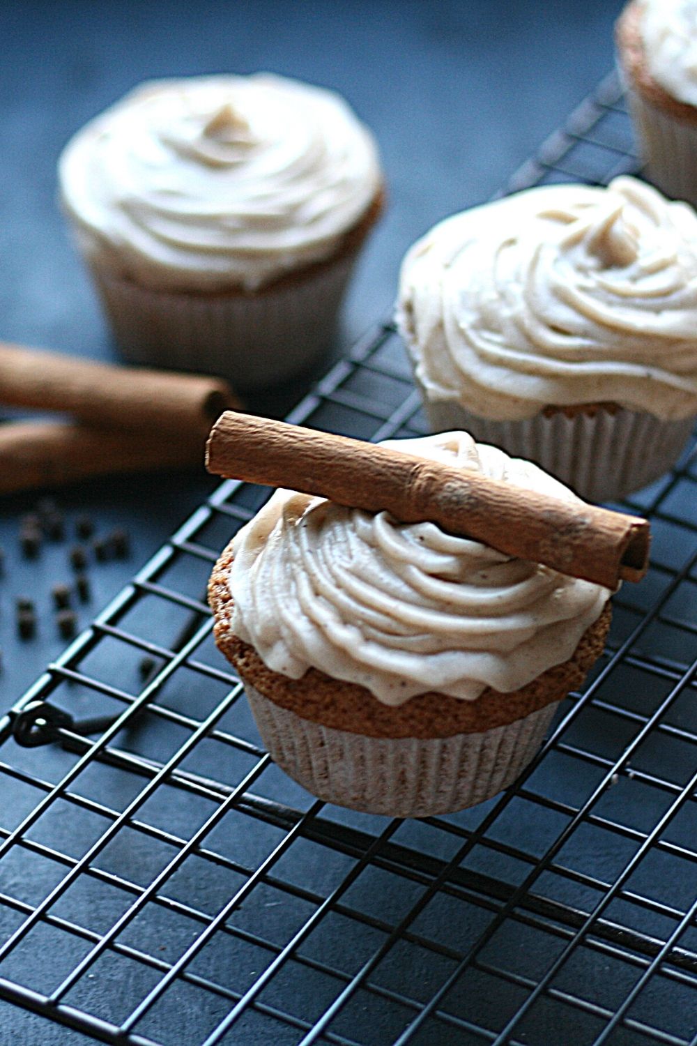 Chai cupcakes with spiced cream cheese frosting decorated with a cinnamon stick, on a rack, with 2 other cupcakes, cinnamon sticks and peppercorns