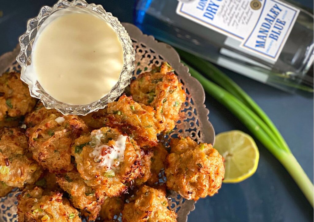 Delicious and simple, warm chicken balls with homemade gin mayonnaise, pictured on a silver tray, with half a lime, a fresh spring onion and a bottle of gin on a dark blue background